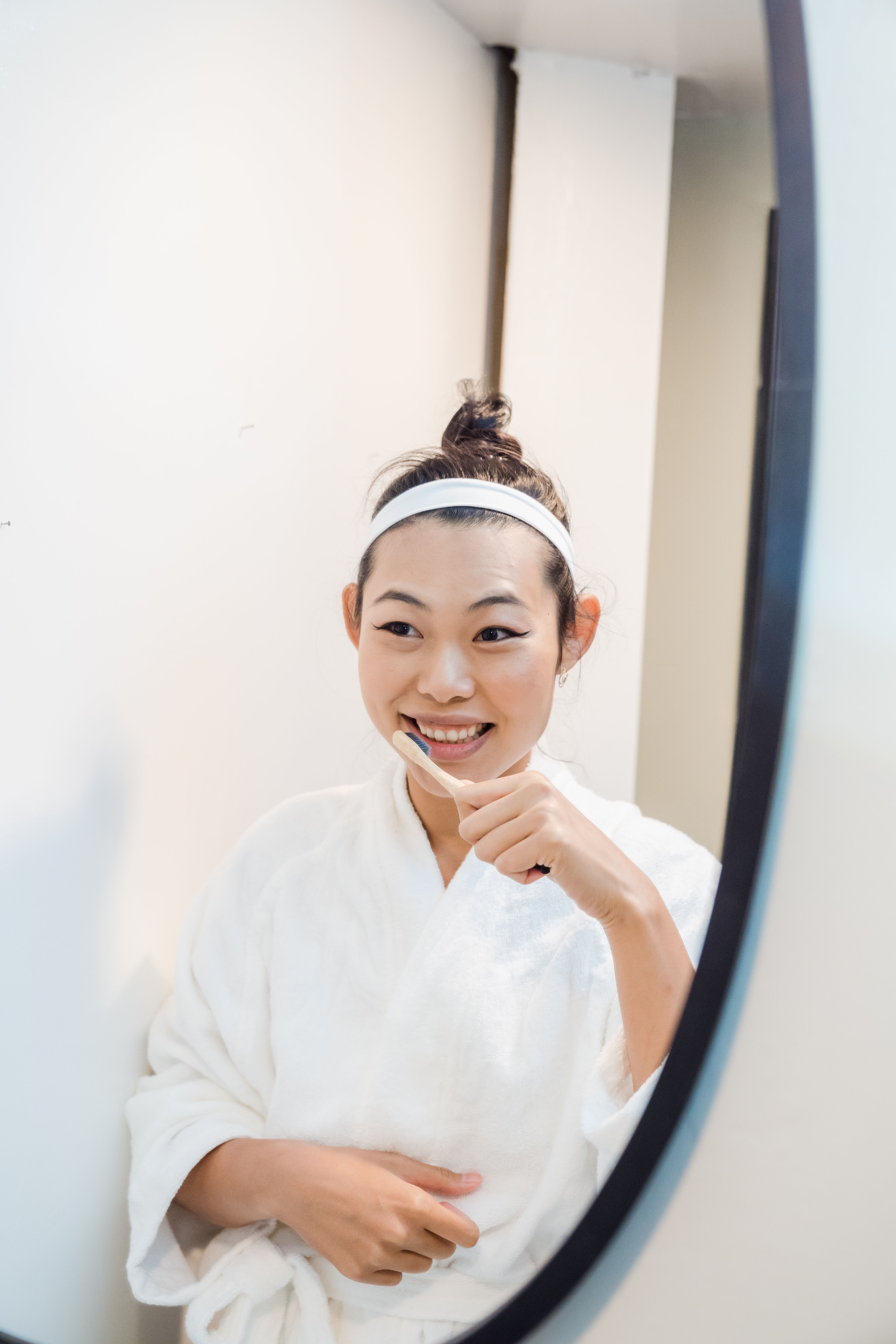 Smiling Asian woman brushing her teeth in the mirror, focusing on dental hygiene and personal care.