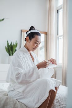 A young woman in a bathrobe sitting on a bed applying a facial mask for skin care.