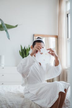 Woman in bathrobe applying facial mask in a bright, serene bedroom. Perfect for skincare and wellness themes.