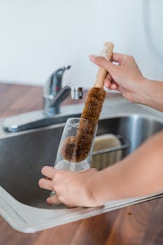 Close-up of hands cleaning a glass with a brush in a kitchen sink.