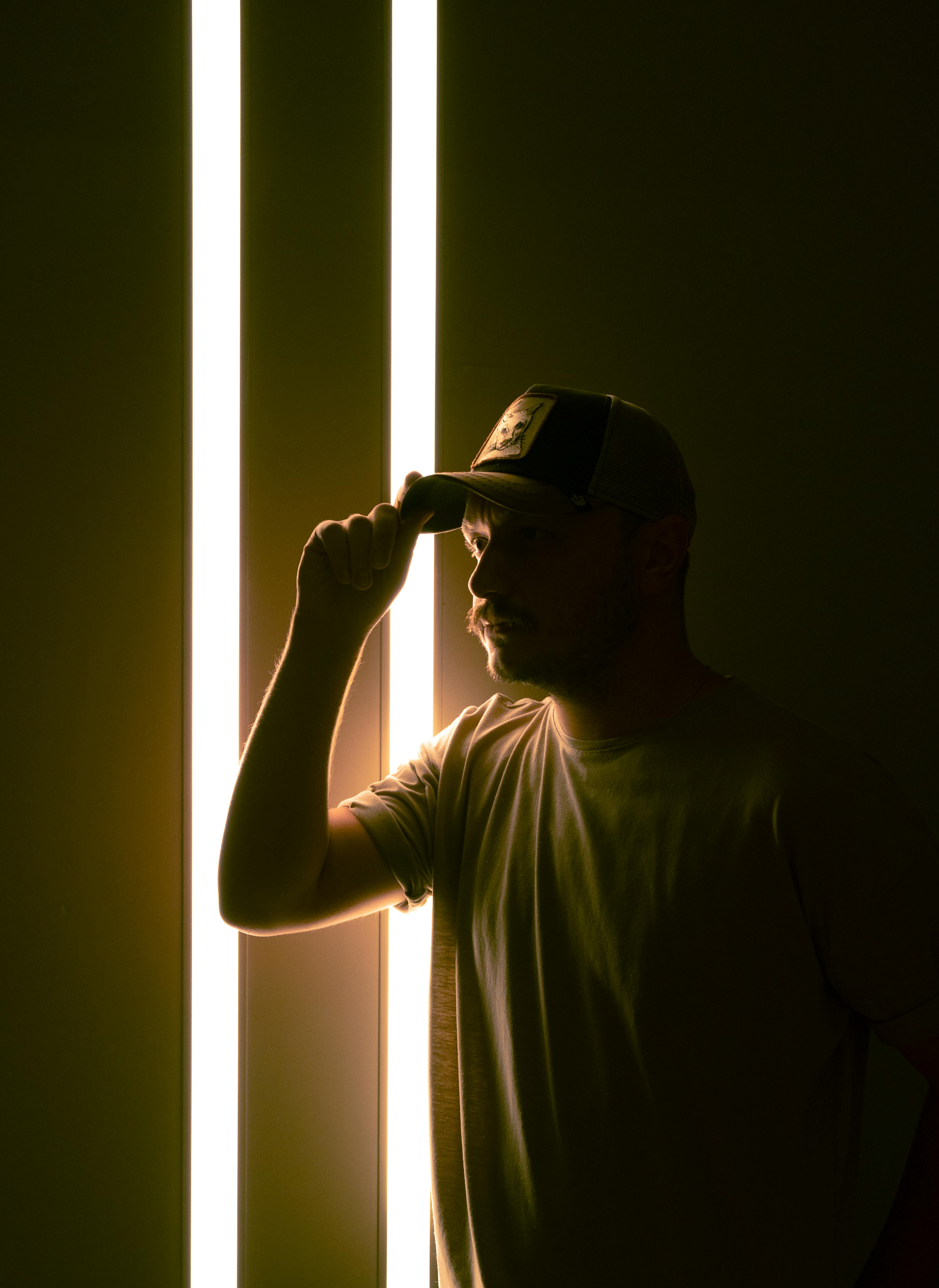 A man stands before vertical neon lights, creating a dramatic silhouette as he tips his cap.