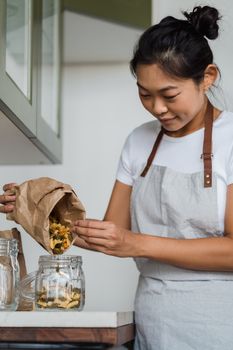 Asian woman carefully transfers uncooked spiral pasta into a glass jar in a modern kitchen setting.
