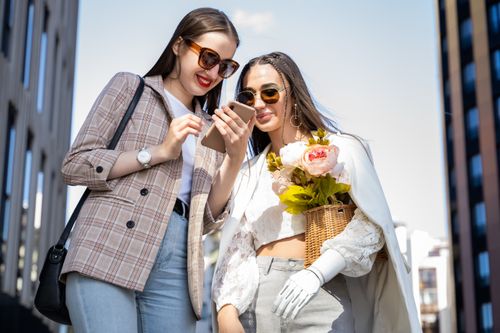 Free Two stylish women enjoy moments in the city with flowers and phones on a sunny day. Stock Photo