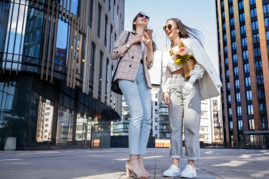 Two fashionable women laughing with a bouquet outdoors in the city, embodying urban style and friendship.