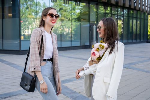 Free Two fashionable women with sunglasses and flowers chatting happily outdoors. Stock Photo