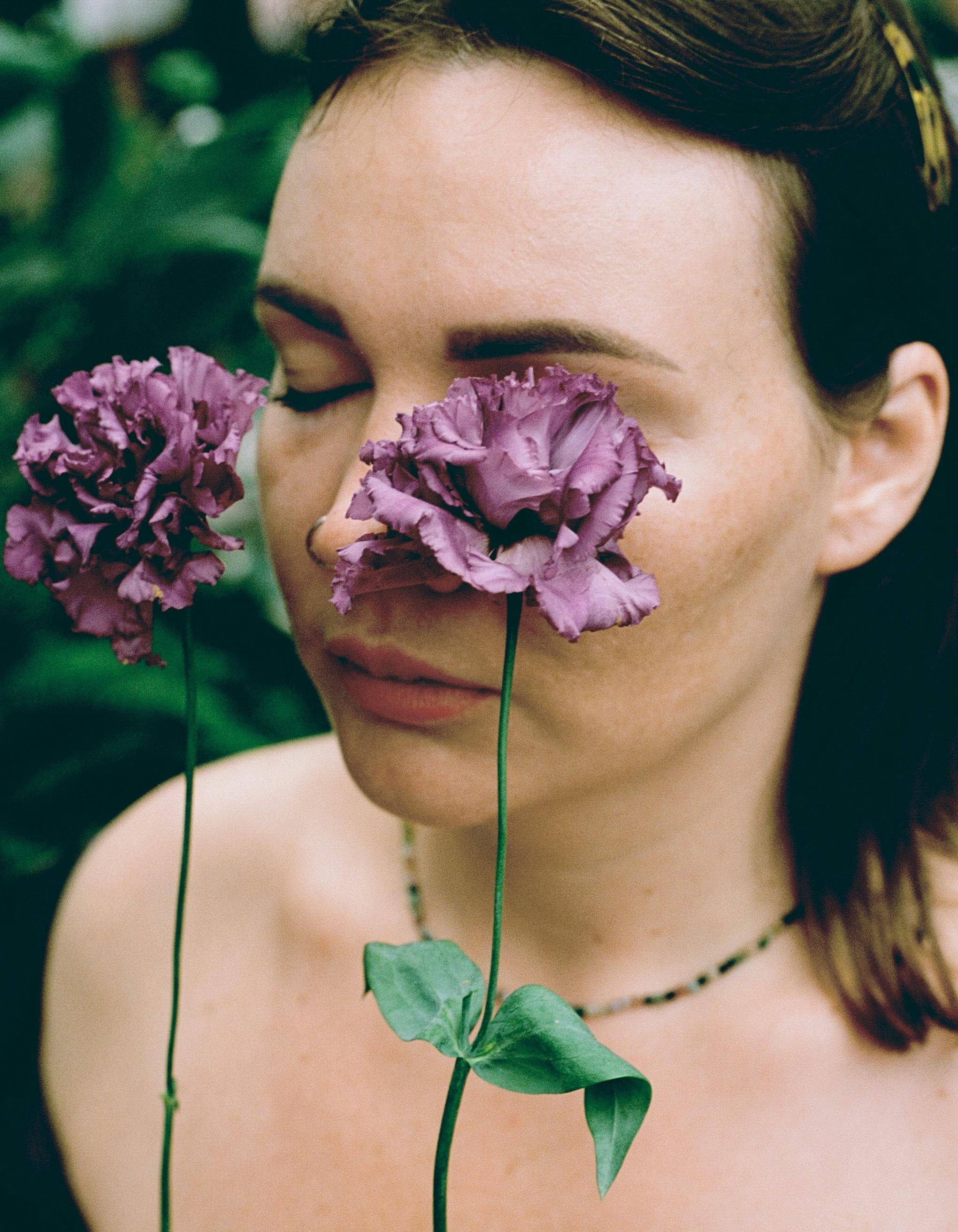 Portrait of a woman with purple flowers in a serene outdoor setting, eyes closed.