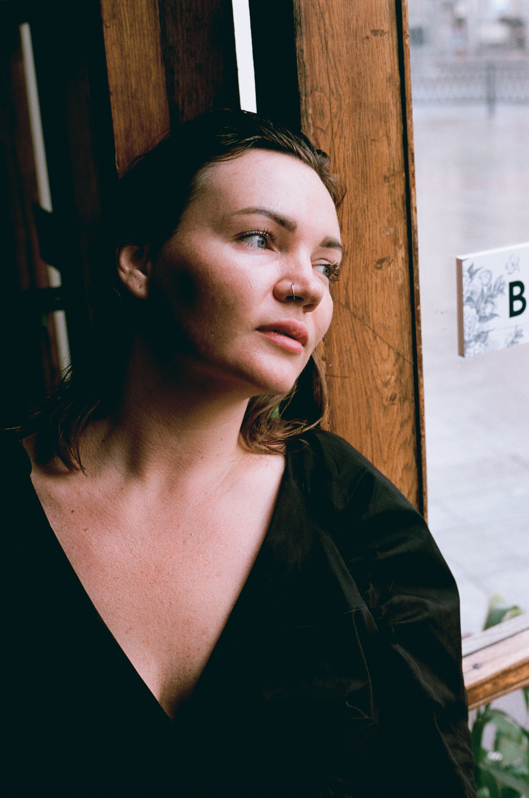 A thoughtful woman leaning against a wooden wall indoors, gazing outside.