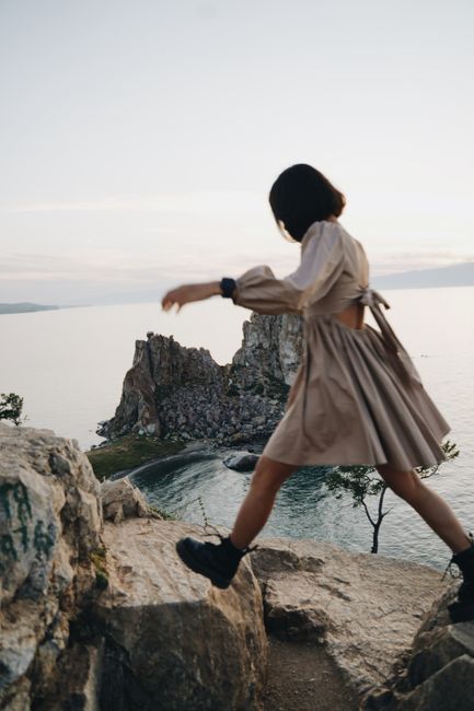 A woman in a beige dress and black boots walks on a rocky cliff by the seaside, capturing adventure and freedom.