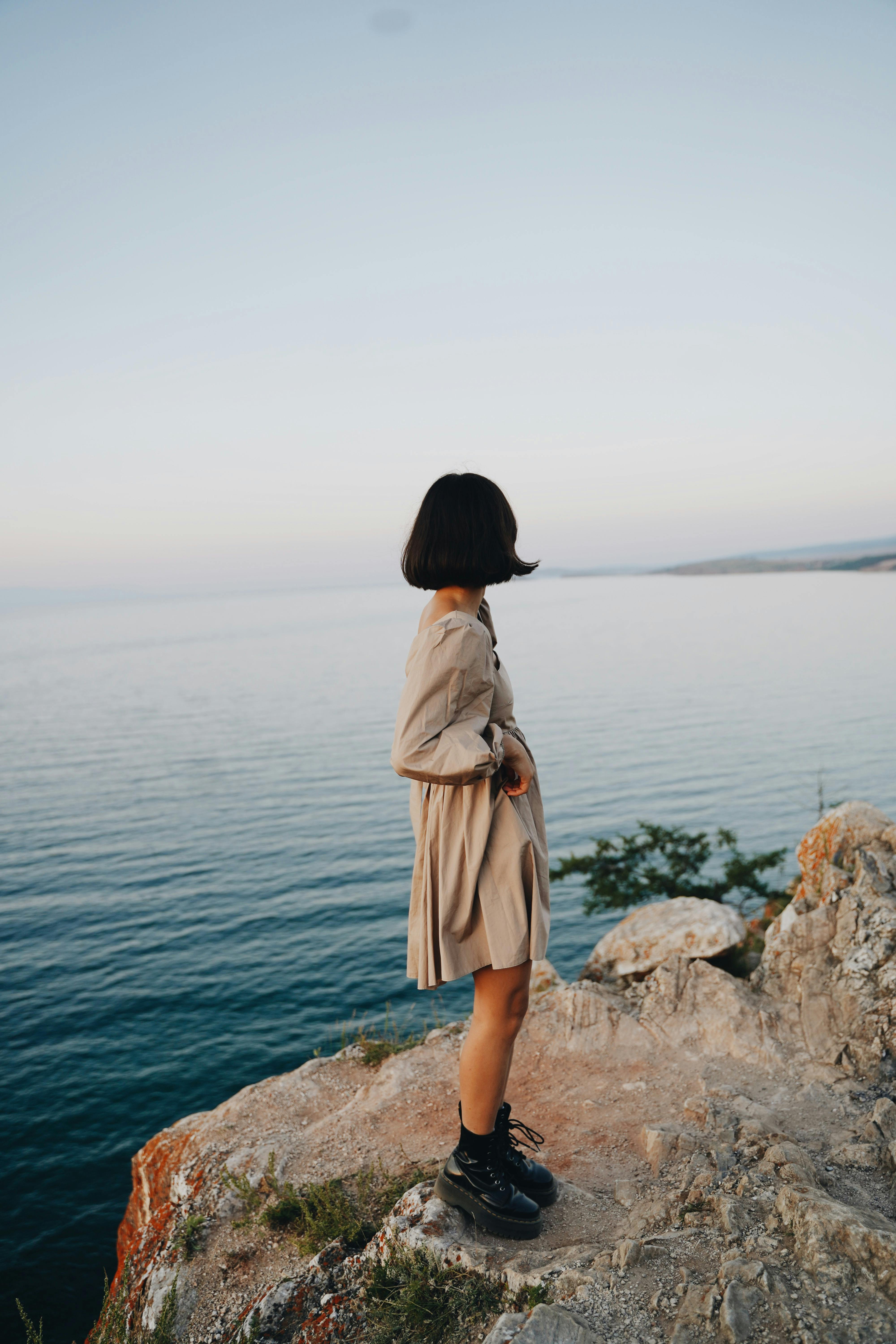 A woman in a brown dress stands poised on a cliff edge, gazing over a serene ocean vista.