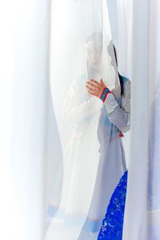 A woman elegantly standing behind a sheer white curtain, wearing traditional clothing with vibrant accents.