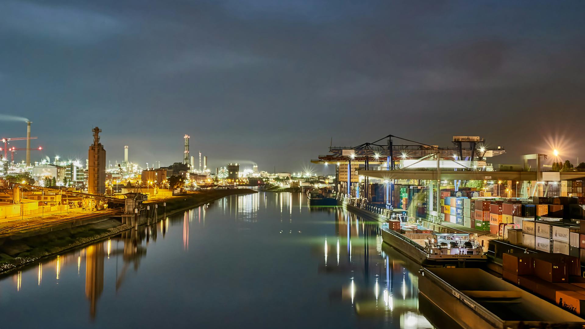 Nighttime view of an illuminated port with shipping containers reflecting on a calm river.