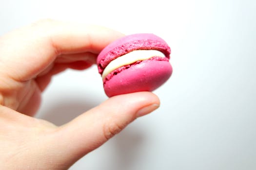 Close-up of a pink macaron held between fingers, showcasing its delicate design.