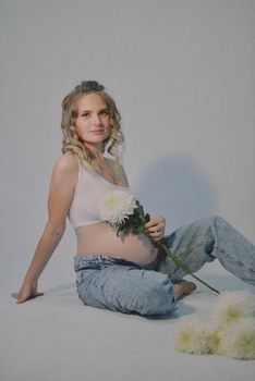 Smiling pregnant woman seated on floor with a white flower, in casual attire.