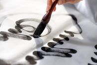 Close-up of a hand using a calligraphy brush to apply black ink on white paper.