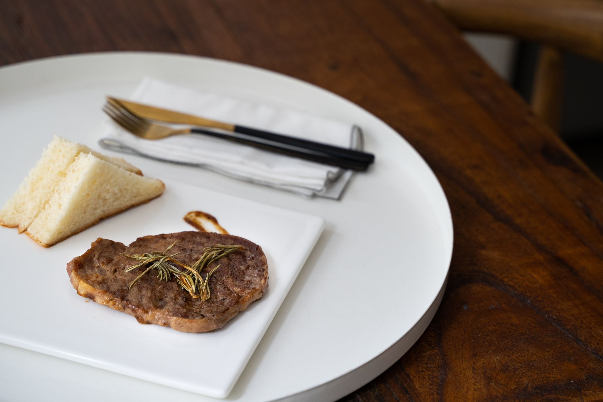 A delectable steak with rosemary lies next to sandwich bread on a minimalist plate, set elegantly with cutlery.