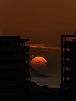 Dramatic sunset over the ocean with city buildings silhouetted against an orange sky.
