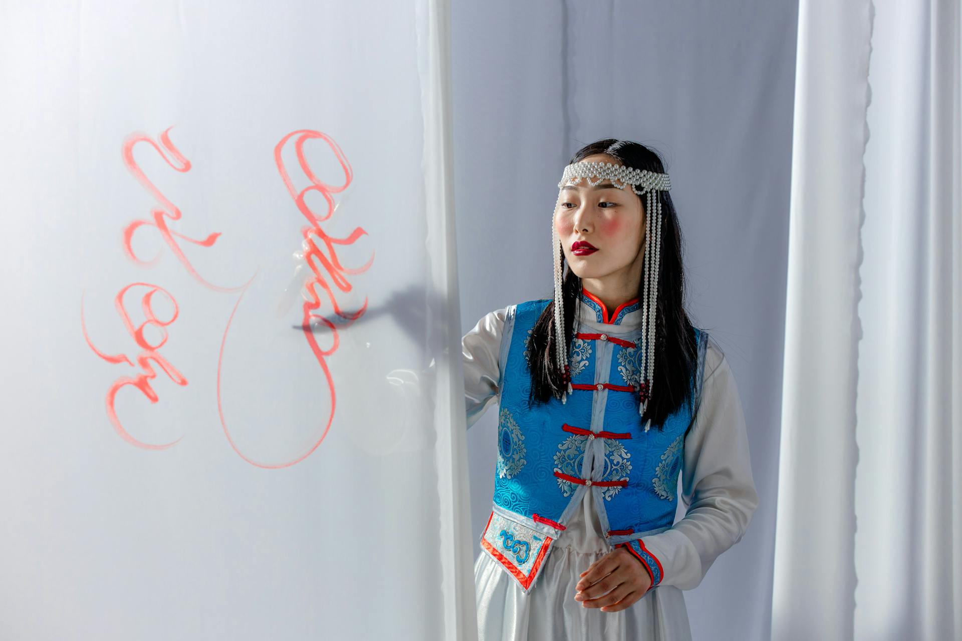 An Asian woman in traditional attire paints red calligraphy on a white curtain indoor setting.