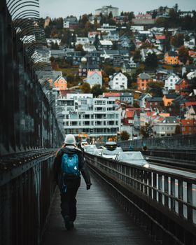 A pedestrian walks on Tromsø Bridge with colorful houses in the background on a cold day.