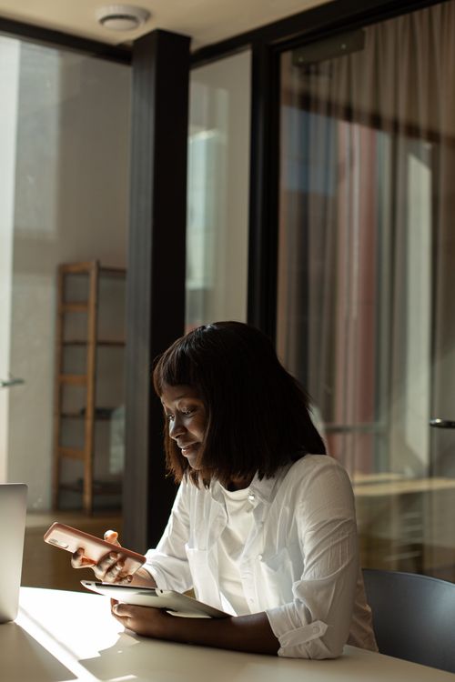 Free African American woman seated in a modern office, engaged with her smartphone at a desk. Stock Photo