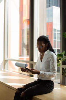 A focused young black woman reading documents while sitting by a window in a modern office.