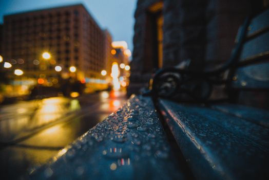 A night cityscape featuring a rain-soaked bench with vibrant blurred city lights in the background.