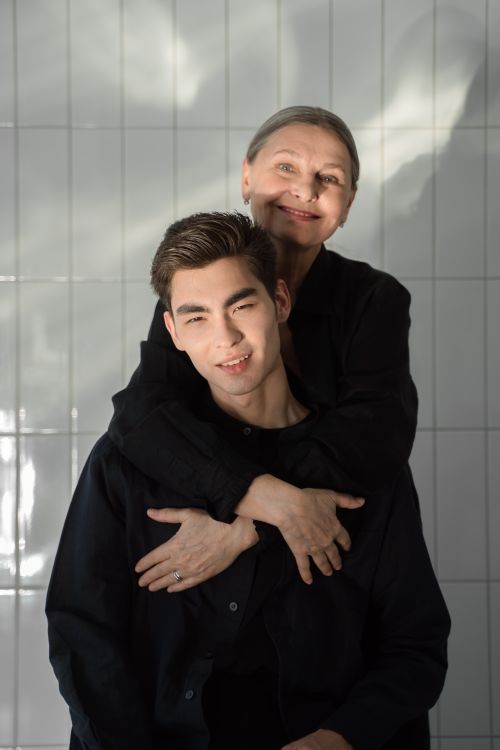 Free An elderly woman lovingly embraces a young man against a tiled wall, both smiling warmly. Stock Photo