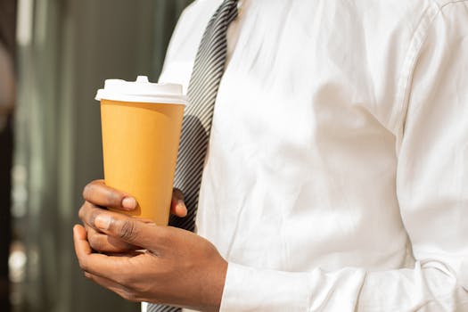 Close-up of a businessman holding a disposable coffee cup outdoors, wearing a tie and dress shirt.