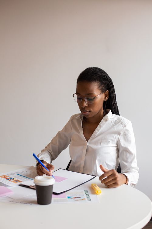 Free Young African woman writing notes at desk with papers and coffee cup. Stock Photo