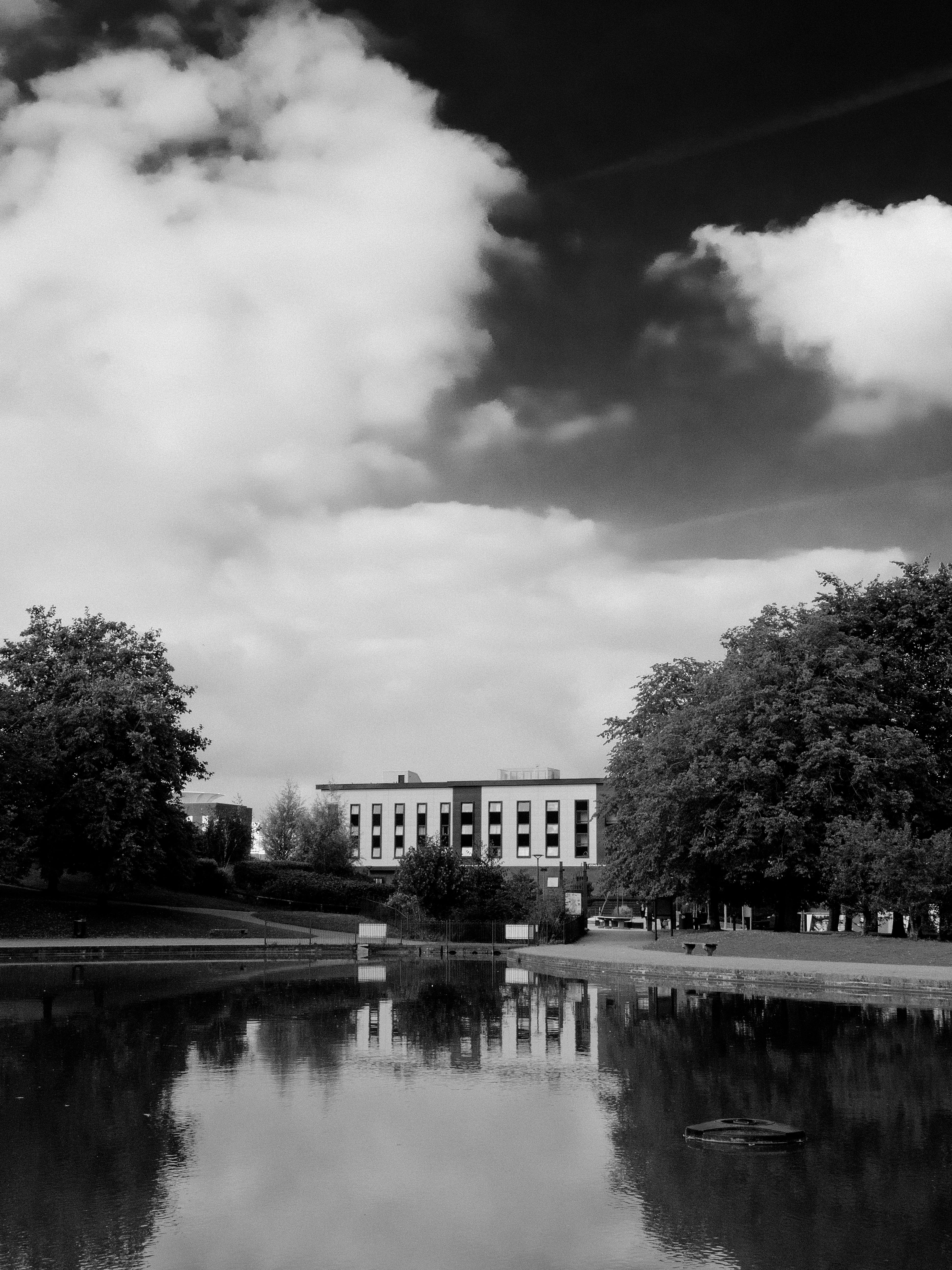 A black and white photo capturing a building reflected in a lake with trees and clouds.