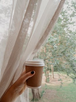 A hand holding a coffee cup near a window with a view of a peaceful park and trees.