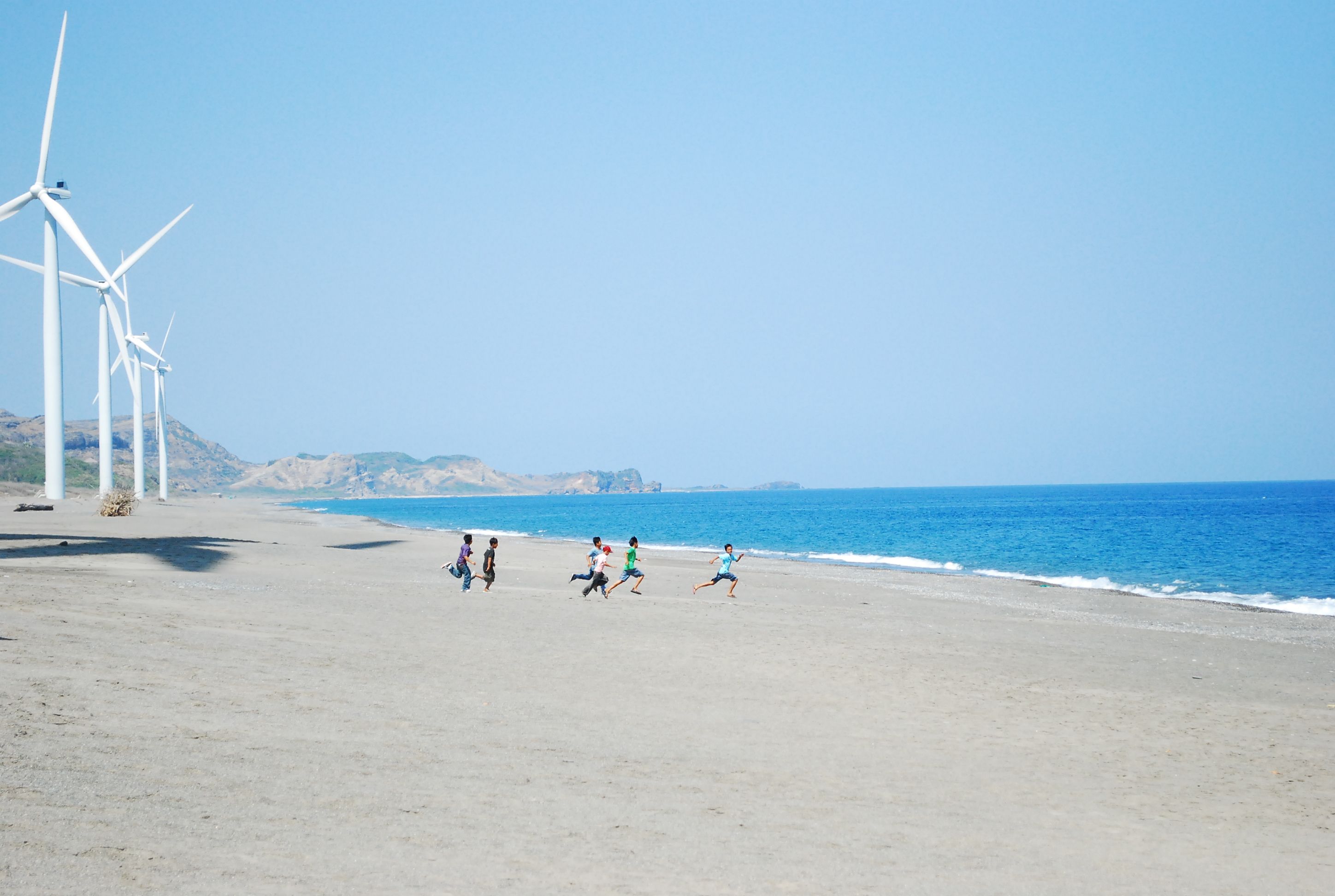 Kids playing and running on a sandy beach with windmills and blue sea in the background.