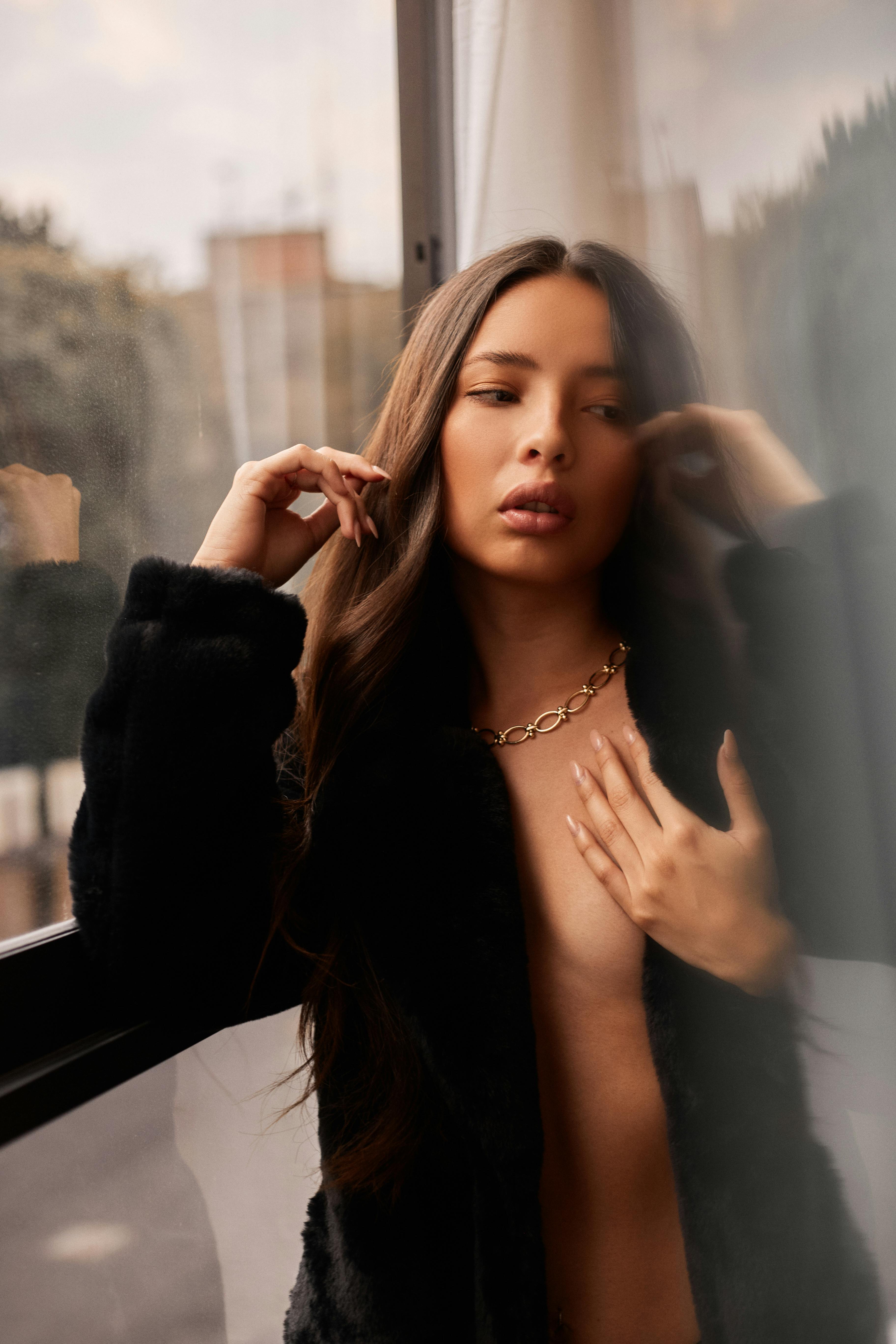 Sophisticated woman in a fur coat poses by the window, exuding style and grace.