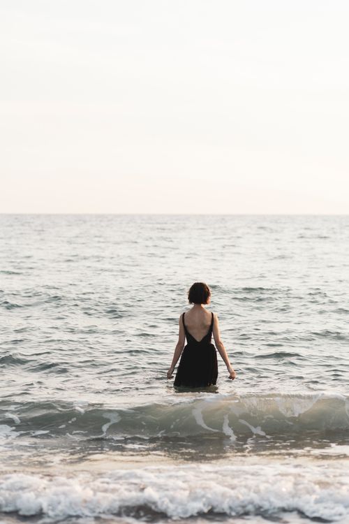 Free Back view of a woman in a black swimsuit wading into the ocean, embracing peaceful solitude. Stock Photo