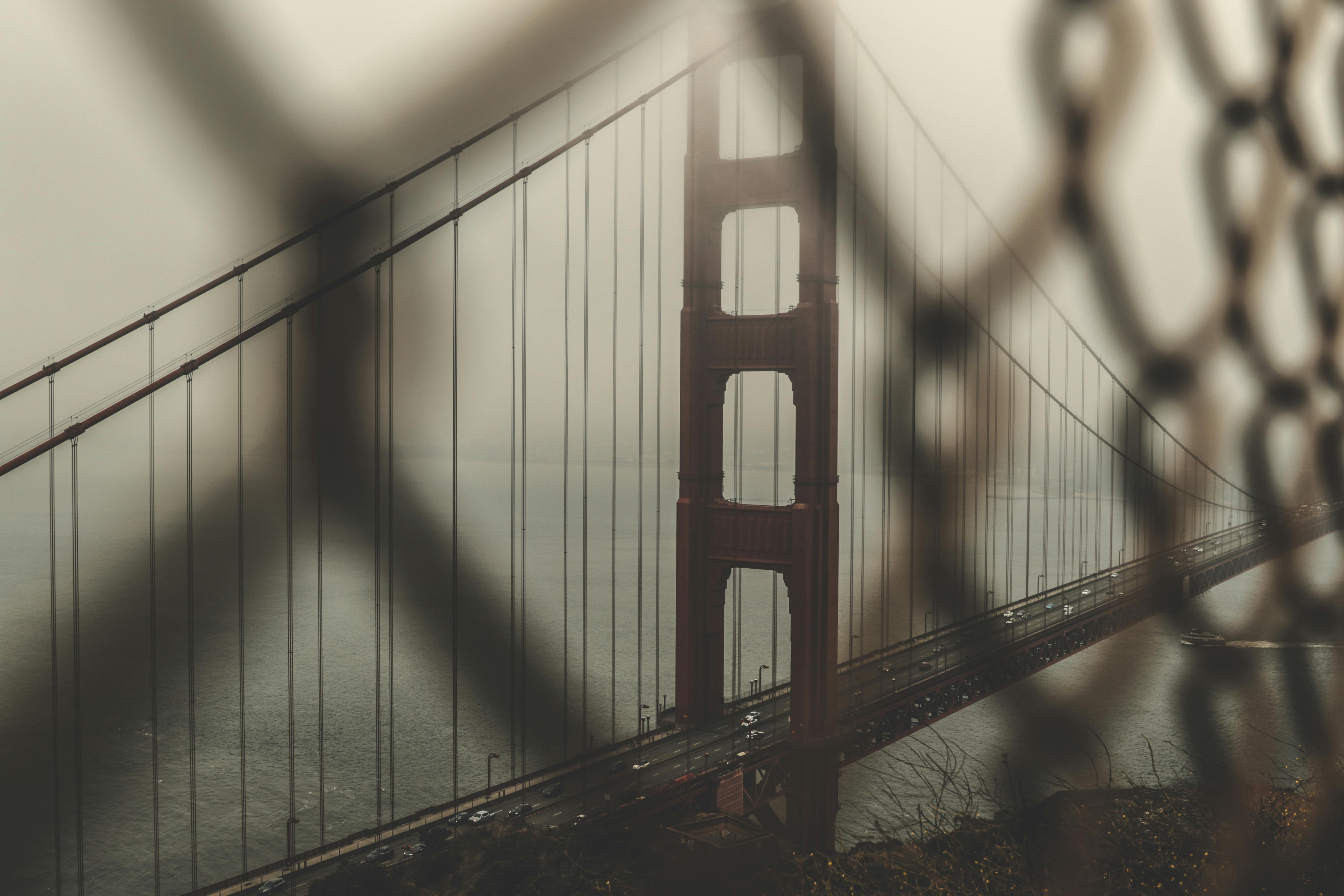 Iconic Golden Gate Bridge seen through a fence, enveloped in moody mist during sunrise.