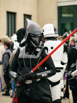 A Star Wars cosplayer in a Sith costume holding a red lightsaber among a crowd outdoors.