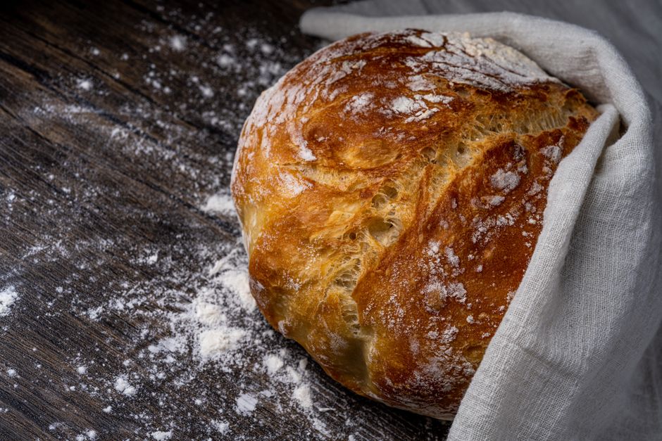 Freshly baked artisan bread loaf dusted with flour on a rustic wooden table, wrapped partly in cloth.
