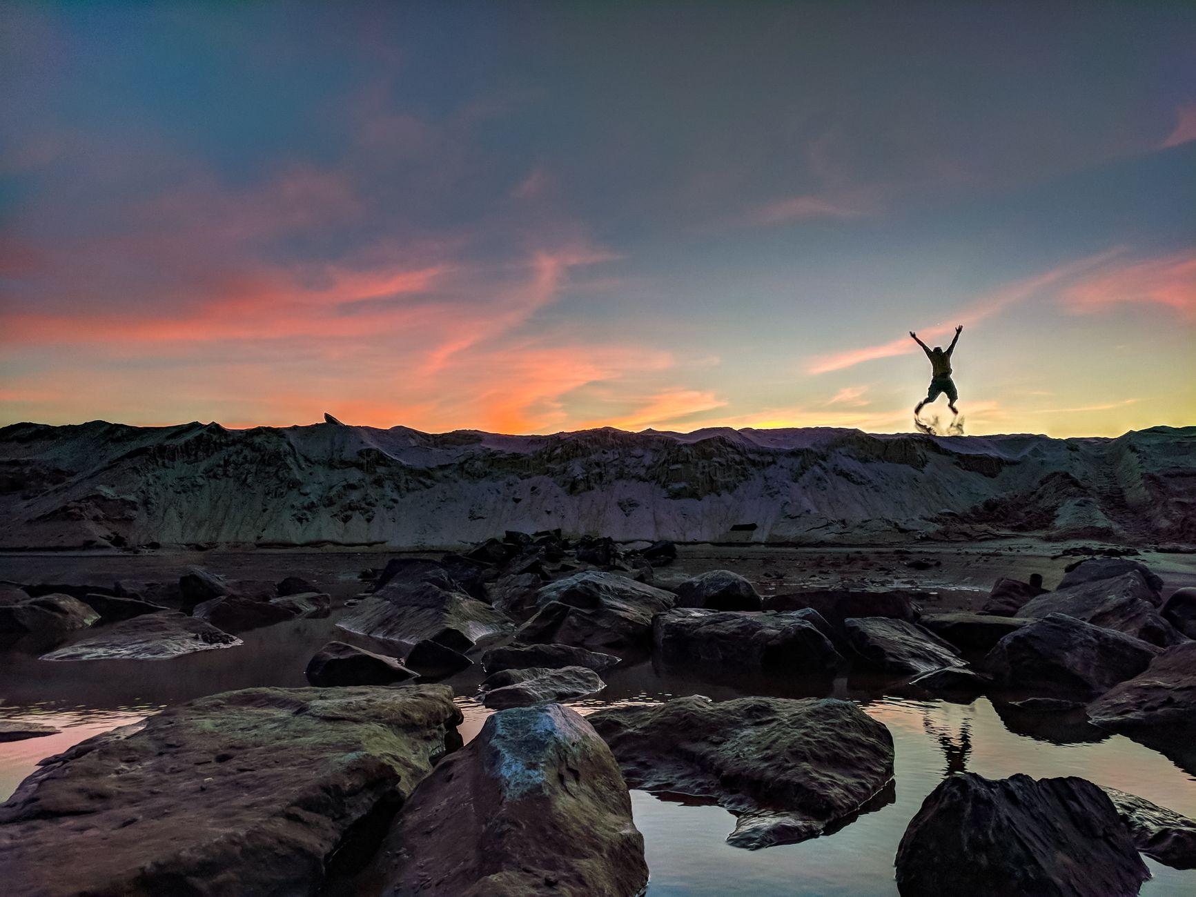 A person silhouetted in mid-air jumping over rocks at sunset. The sky is painted with vibrant colors.