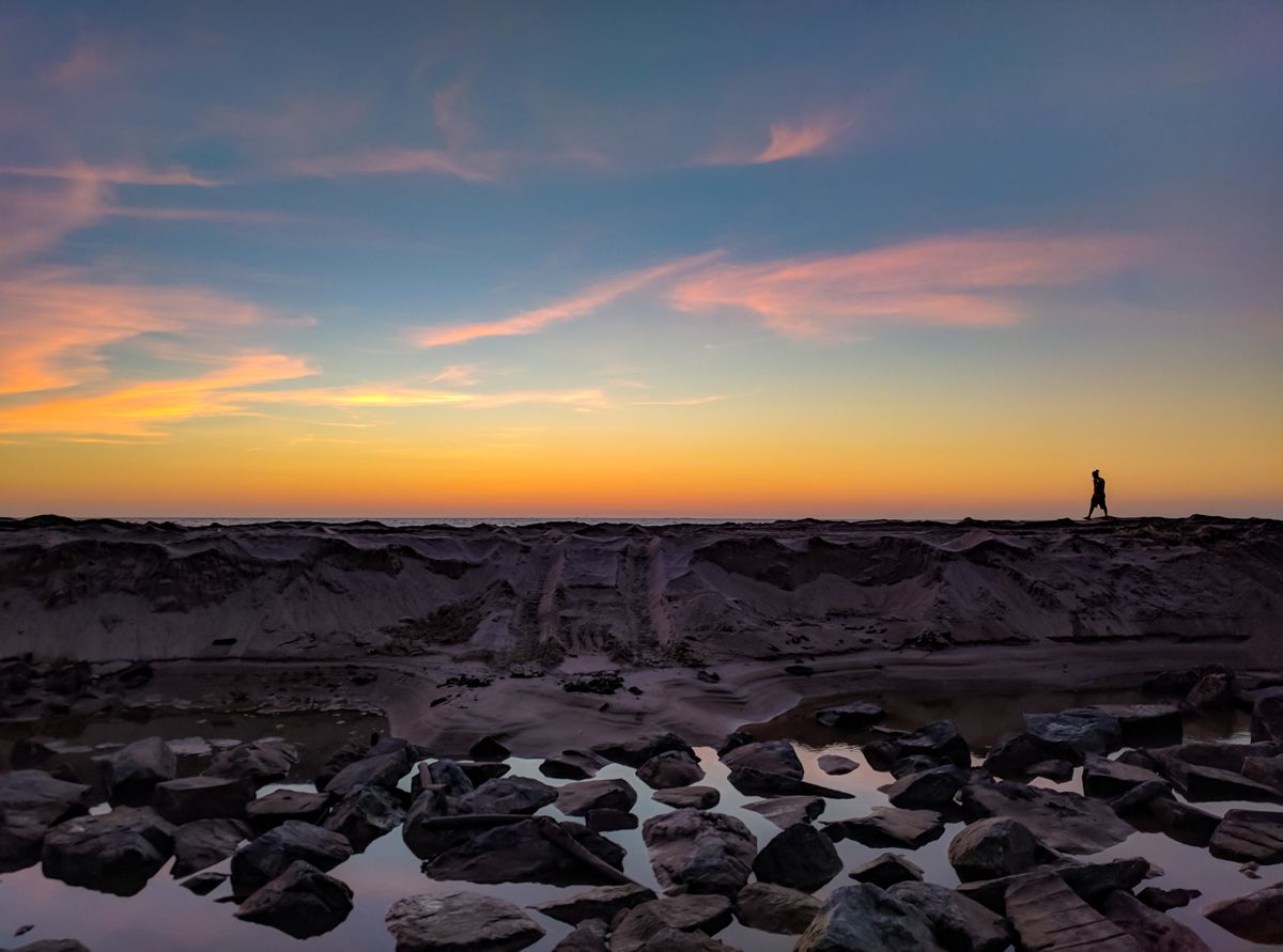 Silhouette of a person walking along a rocky shoreline at sunset, tide pools reflecting orange sky