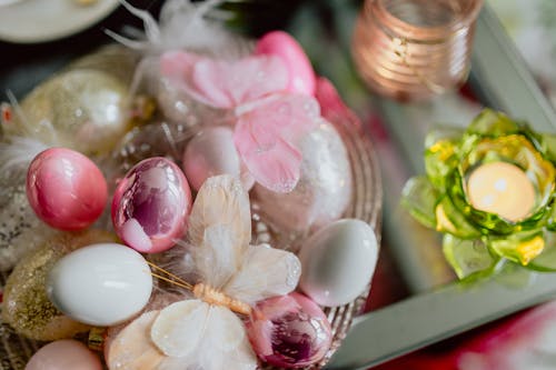 From above of shiny Easter eggs in bowl with decorative artificial butterflies placed on glass table with small candle