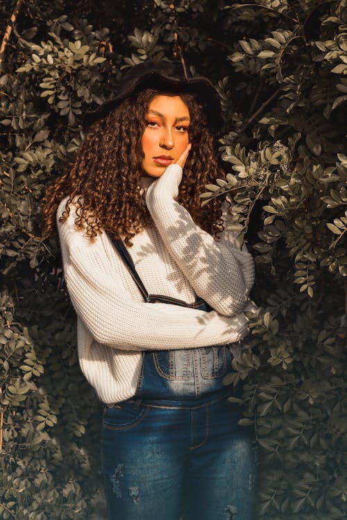 Woman in White Sweater and Denim Jumper Standing Beside Green Leaves