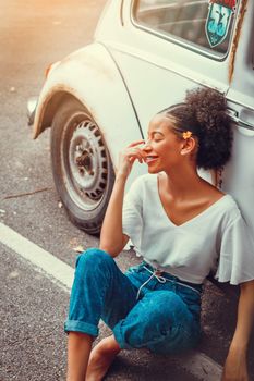 A smiling woman with flower in hair sitting against a parked vintage white car on a sunny day.