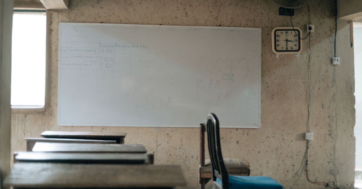 A dimly lit classroom in rural Volta Region with wooden benches and a chalkboard, students sitting on the floor