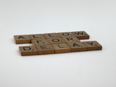 Wooden Scrabble tiles arranged on a white surface spelling 'Allow for Delay'.