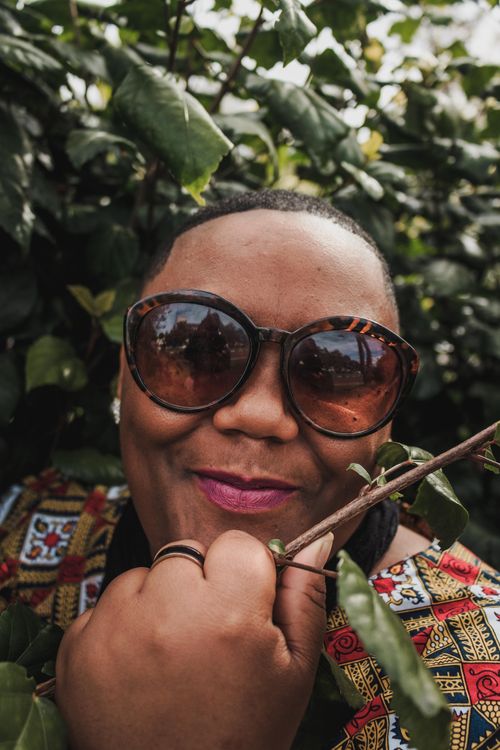 Free Cheerful woman wearing sunglasses amidst green leaves, smiling warmly. Stock Photo