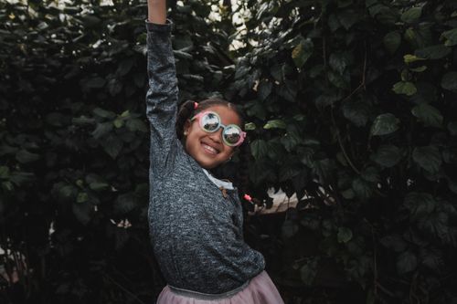 Free Happy girl in pink sunglasses raising her arm in a lush green garden, smiling joyfully. Stock Photo