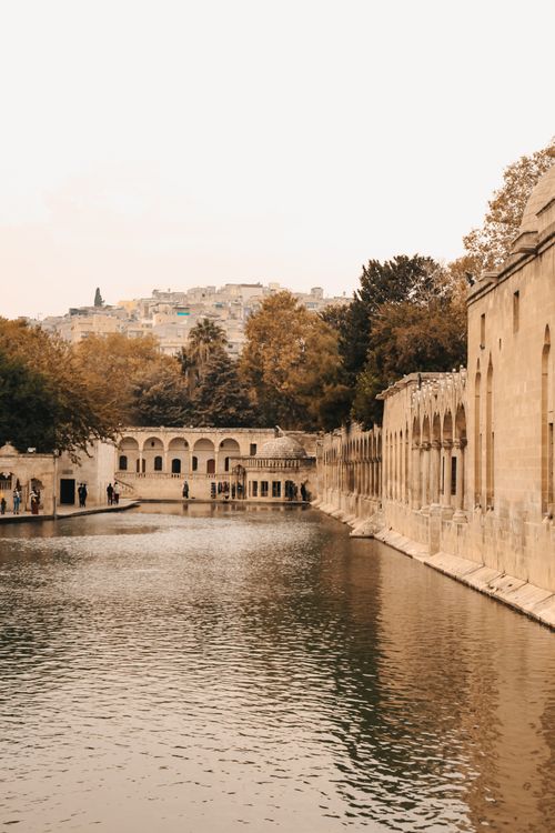 Free A scenic view of ancient stone buildings lining a tranquil pond, framed by lush green trees. Stock Photo