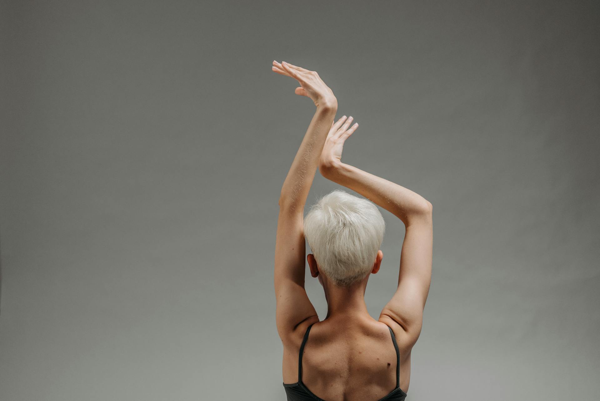 Person with short white hair poses with extended arms against a neutral backdrop.