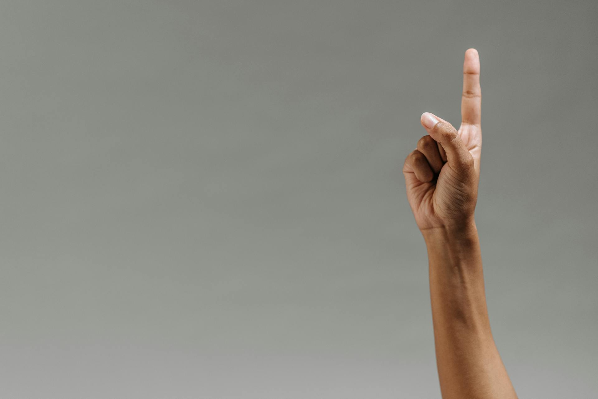 Close-up of a hand raised with the index finger pointing up against a gray background.