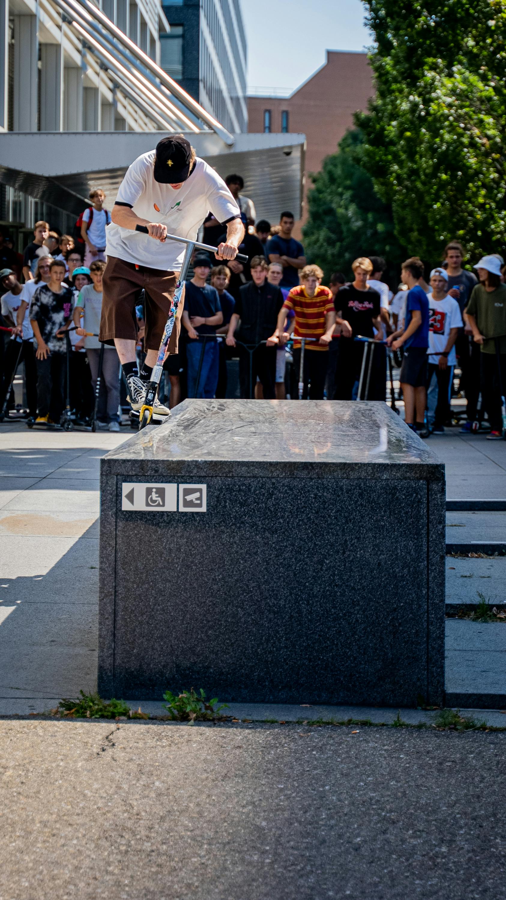 Scooter rider performing a trick at a skatepark with a crowd watching, capturing urban sports culture.