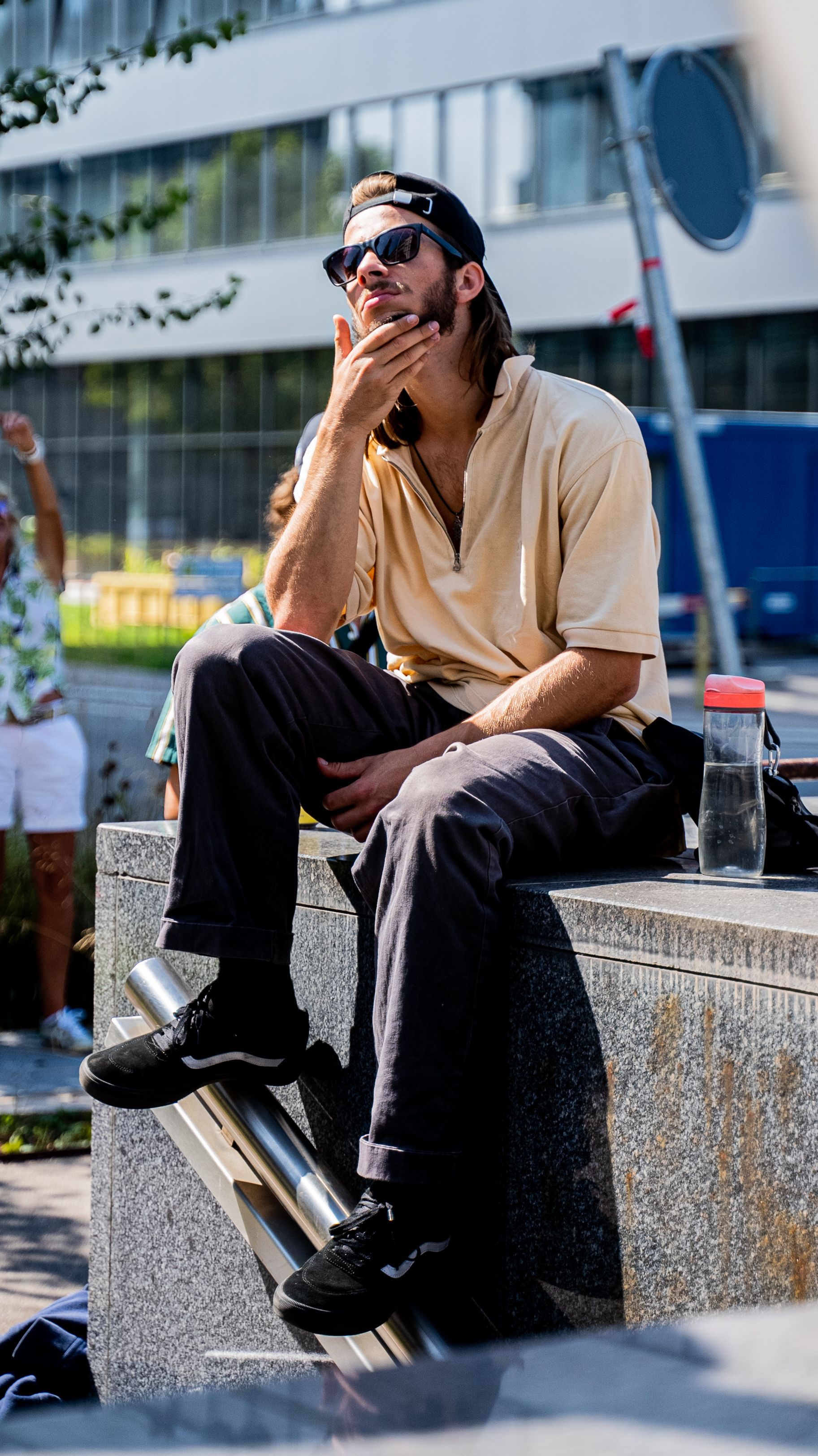 A stylish man in sunglasses sits thoughtfully in an urban setting.